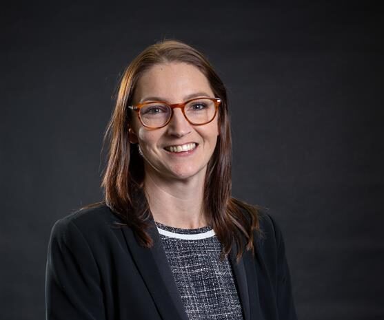 Portrait of Claire Owens, CEO of Alpine Group, smiling and wearing glasses and a dark blazer against a dark background
